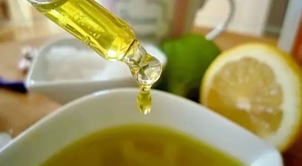 Tropfen eines goldenen Öls fallen aus einer Pipette in eine Schale. Zitronen und Limetten im Hintergrund deuten auf natürliche Inhaltsstoffe für Kosmetik hin, wie z.B. Zitronensäure.