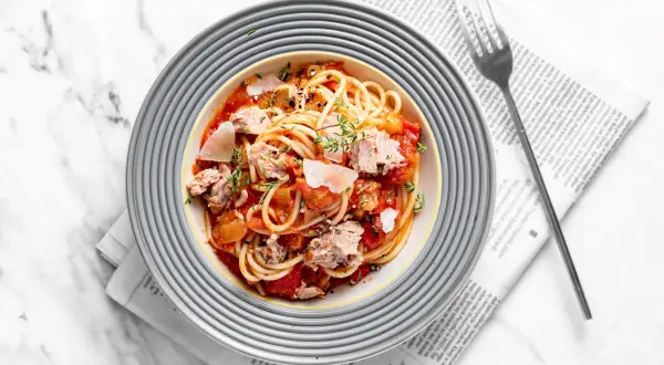 Pyszny makaron z tuńczykiem w sosie pomidorowym, posypany parmezanem i ziołami, podany na talerzu na tle gazety.