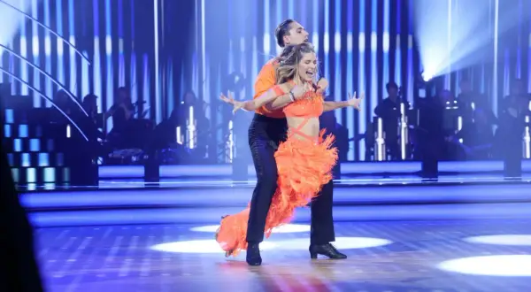 Tancerze w pomarańczowych strojach wykonują dynamiczny big dance. W tle orkiestra, a na scenie migoczą niebieskie światła, jakby sypały się pieniążki.