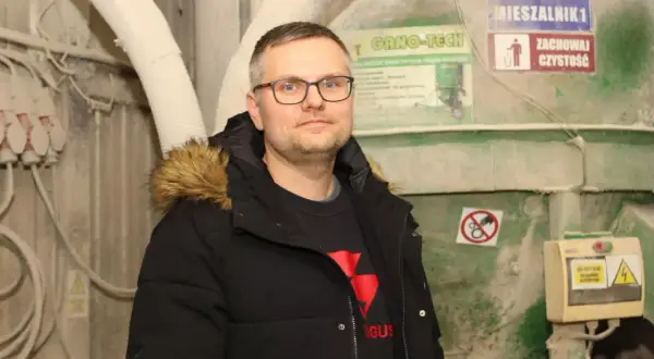 Mężczyzna w okularach i czarnej kurtce z futrzanym kapturem stoi na tle przemysłowych maszyn.