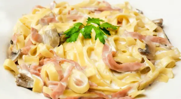 Tagliatelle z szynką i pieczarkami w kremowym sosie, posypane natką pietruszki. Pyszny makaron z mozzarellą w tle.