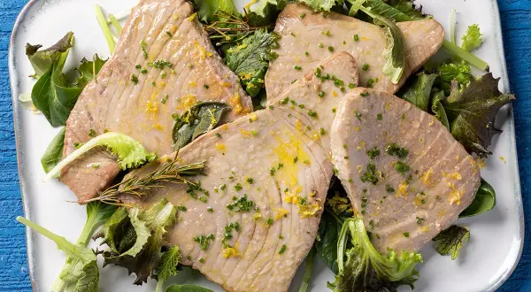 Fette di tonno al forno, dorate e succulente, servite su un letto di insalata fresca con scorza di limone e erbe aromatiche.