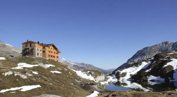 Das Pfitscher Joch Haus thront auf einem Hügel neben einem Bergsee. Die Alpenlandschaft ist von Schnee und braunem Gras bedeckt.