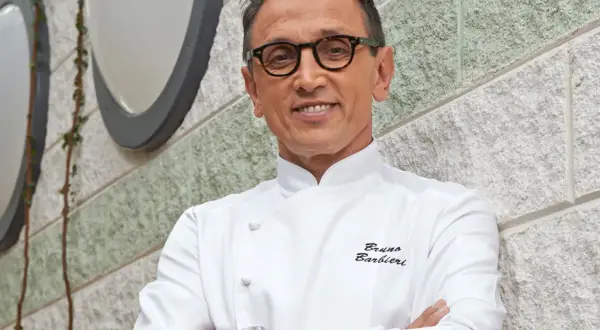 Bruno Barbieri, chef stellato Michelin, sorride con le braccia conserte davanti a un muro di pietra.