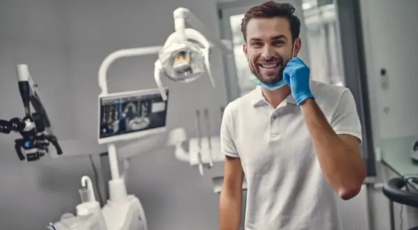 Uśmiechnięty dentysta w gabinecie, gotowy do pracy. Zastanawiasz się, ile trwa plombowanie zęba?