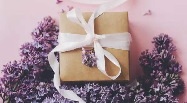 Ein Geschenkpaket mit weißem Band, umgeben von lila Flieder. Ein schönes Zeichen zum 13. Hochzeitstag.