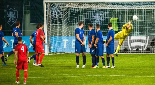 Piłkarze w niebieskich strojach czekają na wznowienie gry. Bramkarz w żółtym stroju broni strzał. Czas na przerwę w meczu?
