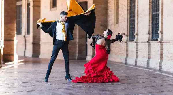 Pareja bailando flamenco, él con capa negra y amarilla, ella con vestido rojo voluminoso. La imagen evoca la diferencia entre flamenco y sevillanas.