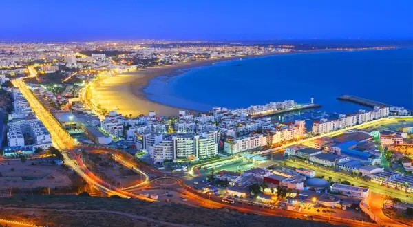 Agadir, Marocko, upplyst vid skymningen. En livlig stad vid havet med en strandpromenad, en marina och ljusspår från trafiken.