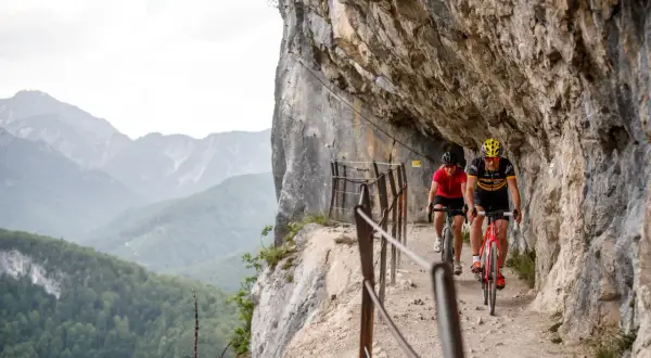 Dwóch rowerzystów pokonuje malownicze trasy rowerowe w Austrii, jadąc wąską ścieżką przy skalnej ścianie z widokiem na góry.