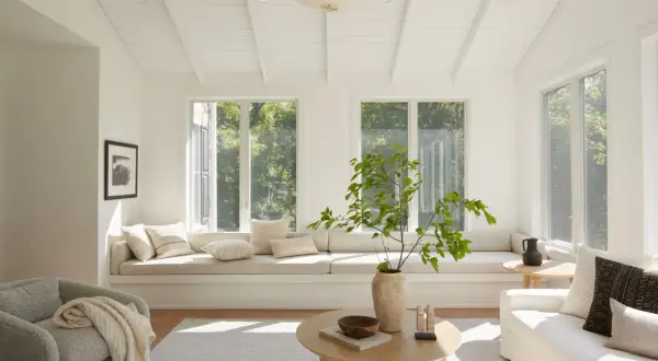 A bright living room with neutral interior design, featuring a large window seat, a textured armchair, and a light wood coffee table.
