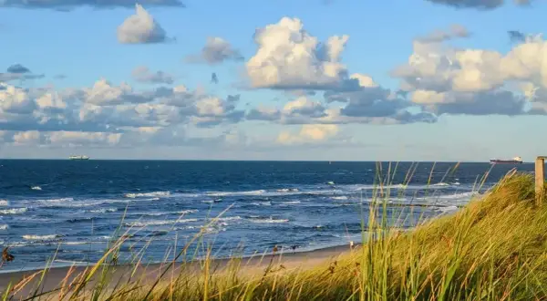 Piękny widok na morze z piaszczystą plażą i wydmami porośniętymi trawą. Na horyzoncie statki, a na niebie chmury. Dźwirzyno atrakcje to idealne miejsce na wypoczynek.