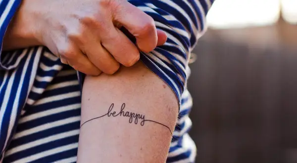 Un brazo con una camiseta a rayas azules y blancas muestra un delicado tatuaje de caligrafía que dice "be happy", una forma de arte para transformar cicatrices.