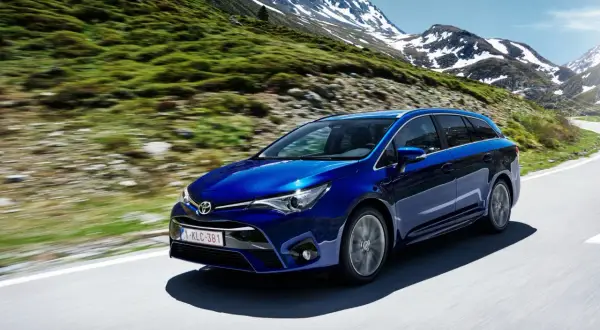 Niebieska Toyota Avensis T27 1.8 benzyna przemierza górską drogę, otoczona zielonymi zboczami i ośnieżonymi szczytami.