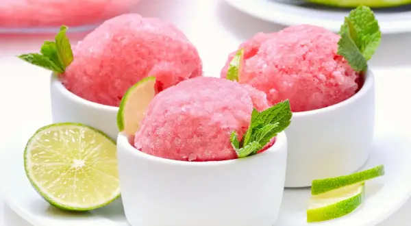 Orzeźwiający sorbet z arbuza w białych miseczkach, udekorowany listkami mięty i plasterkami limonki. Idealny na upalne dni.