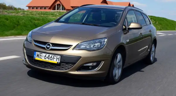 Brązowy Opel Astra J automat w wersji kombi jedzie po drodze. W tle widać domy z czerwonymi dachami.