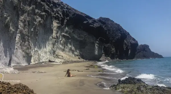 Una mujer descansa en la arena de la cala carbon, con imponentes acantilados de roca volcánica a su espalda.