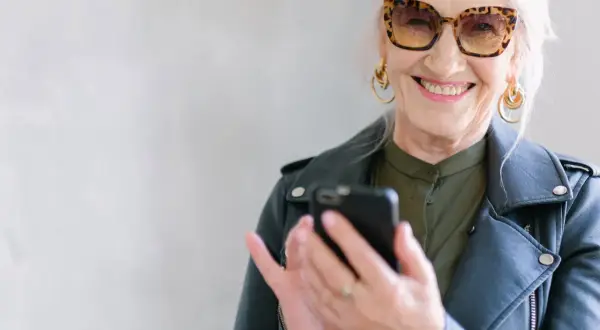 Uśmiechnięta starsza pani w okularach w panterkę sprawdza telefon. Może szuka informacji o kodeksie pracy i odprawie emerytalnej.