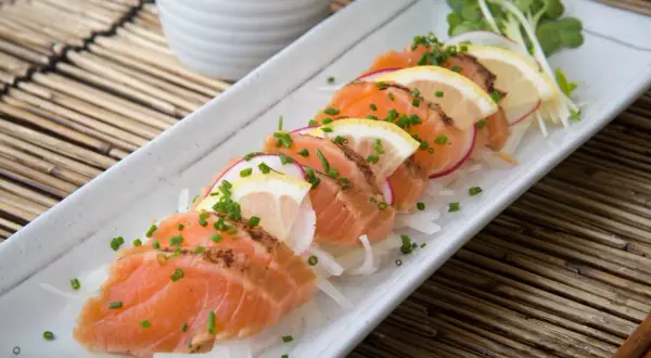 Tataki de salmón fresco, finamente cortado y adornado con rodajas de limón, rábano y cebollino, servido sobre una cama de finas hebras de cebolla.
