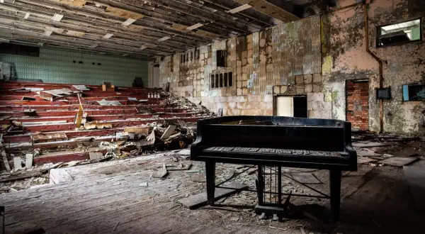 Un piano noir trône au milieu d'une salle délabrée, vestiges de maisons abandonnées. Les sièges rouges sont brisés, le sol jonché de débris.
