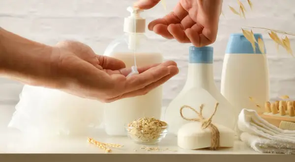 Hands dispensing lotion from a pump bottle into a palm, with oat flakes, soap, and bottles in the background.