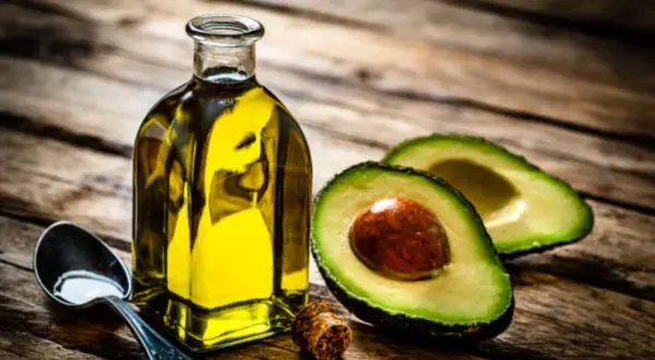 Botella de aceite de aguacate para la piel, junto a un aguacate cortado y una cuchara con aceite.