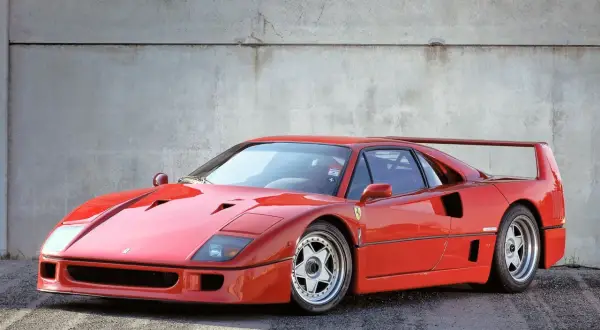 Ikoniczny czerwony Ferrari F40 z charakterystycznym skrzydłem, zaparkowany na tle betonowej ściany.