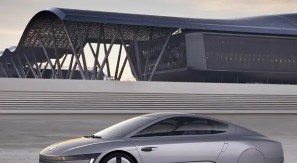Srebrny, futurystyczny vw xl1 z charakterystycznym dachem i nowoczesnym budynkiem w tle.