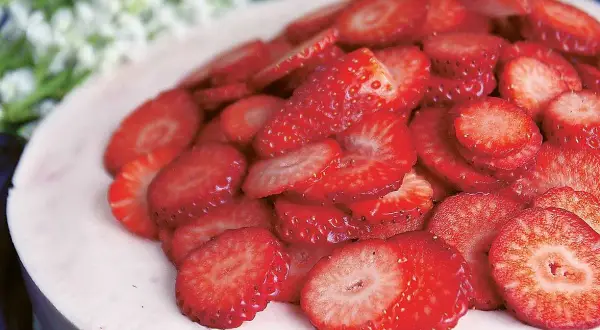 En läcker jordgubbsmousse tårta, toppad med färska jordgubbsskivor och vita blommor i bakgrunden.