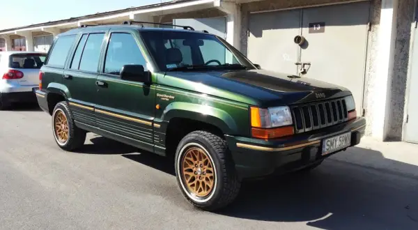 Zielony Jeep Grand Cherokee Limited z lat 90. na złotych felgach, zaparkowany przed garażami.