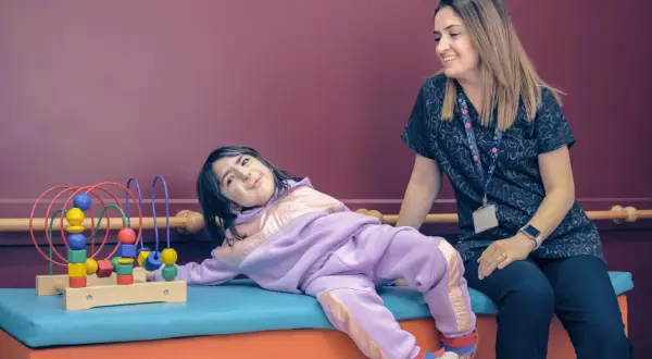 Uśmiechnięta terapeutka patrzy na dziecko bawiące się zabawką terapeutyczną.