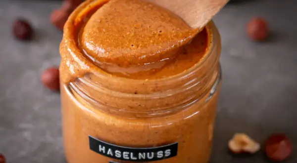 Cremiges Haselnussmus im Glas, bereit für leckere Rezepte mit Haselnussmus. Ein Holzlöffel schöpft die köstliche Masse.