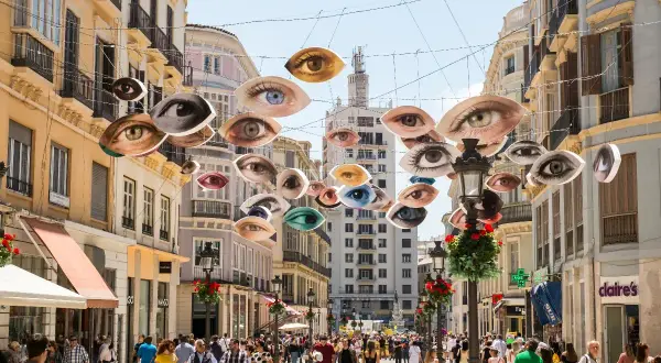 Ojos flotantes decoran una calle famosa de Málaga, observando a la multitud que pasea entre edificios históricos.