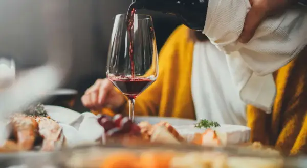 Rödvinet hälls upp i ett glas vid en middagsbjudning. En person i gul tröja sitter bakom. Frågan om hur mycket vin per person är relevant vid sådana tillfällen.