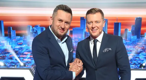 Dwóch mężczyzn w garniturach ściska sobie dłonie na tle scenografii z miejskim krajobrazem, gdzie nagrywane jest koło fortuny.