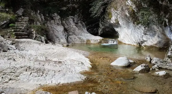 Sentiero di pietra conduce a un ruscello cristallino con piccole cascate di piovere, incastonato tra rocce bianche e vegetazione.