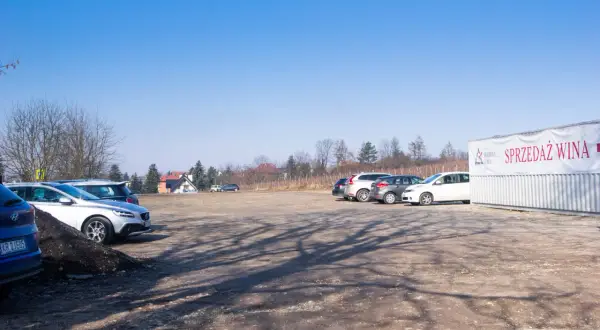 Parking przy Kopcu Piłsudskiego. Kilka samochodów stoi na placu, w tle budynki i drzewa. Na kontenerze widoczny napis "SPRZEDAŻ WINA".