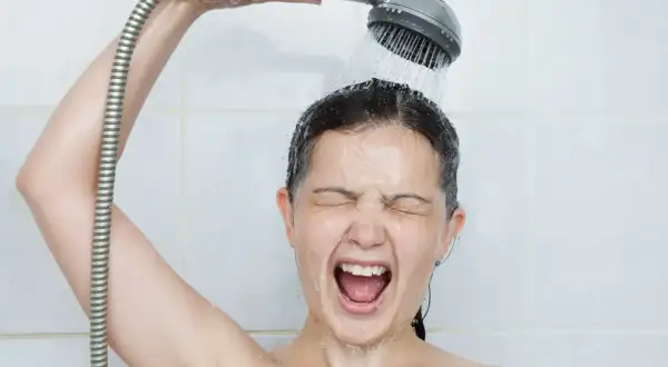 Une femme crie sous une douche froide, visiblement contrariée par un problème d'eau chaude.
