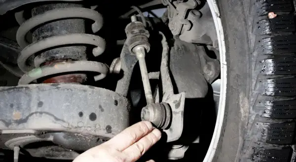 Dłoń wskazuje na zużyty stabilizator w zawieszeniu samochodu, obok sprężyny i opony.