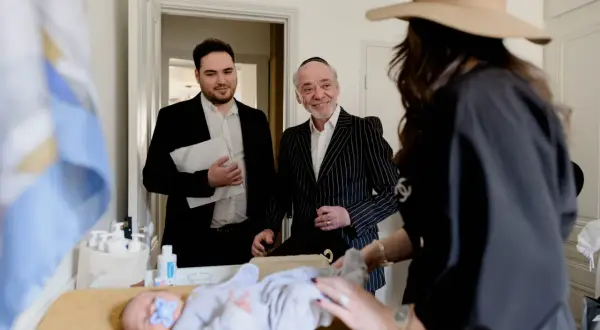 Un bébé est allongé sur une table, une tétine bleue dans la bouche. Des hommes en costume, dont un portant une kippa, observent le déroulement de la circoncision juive.