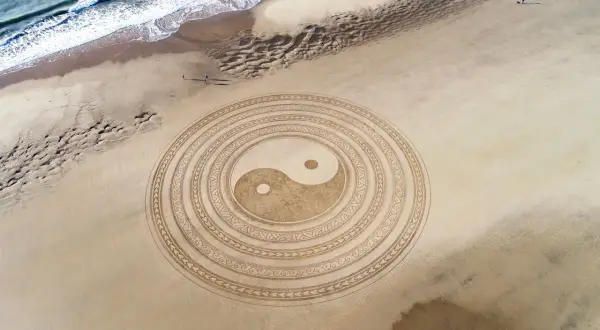 Símbolo Yin Yang en la arena, representando la energía masculina y femenina en equilibrio. El mar y la playa completan la escena.