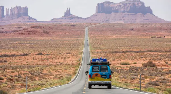 Błękitny van z bagażem na dachu jedzie pustynną drogą w USA. Monument Valley w tle. To część ich wycieczki do USA.