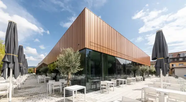 Moderne Architektur der Inselhalle Lindau mit kupferfarbener Fassade, Glasfront und vielen leeren Tischen und Stühlen auf einer gepflasterten Terrasse.