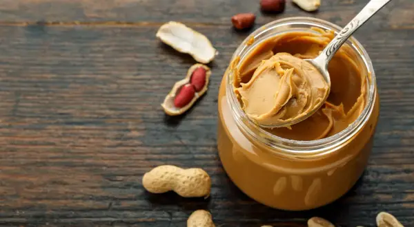 Ein Glas cremige Erdnussbutter mit einer Löffelspitze voll. Nüsse und Schalen liegen verstreut. Perfekt für den Muskelaufbau!