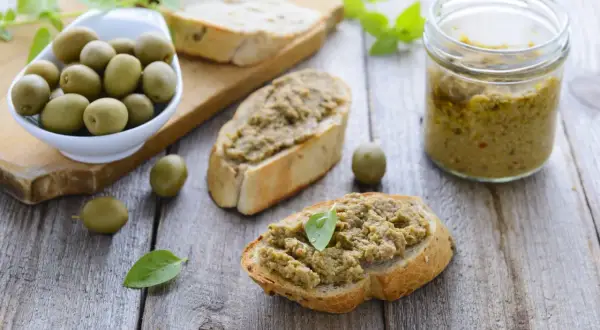 Bruschette con crema di olive, un vasetto di crema di olive e olive verdi su un tagliere di legno.