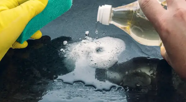 Mano con guanto giallo pulisce con spugna. Aceto sgrassa la superficie, creando schiuma.