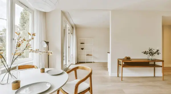 Helles Esszimmer mit weißem Tisch, Holzsessel und Blumen. Ein Regal und ein Beistelltisch mit Deko unterstreichen den "weniger ist mehr lebensstil".