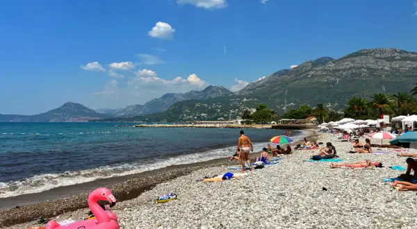 Słoneczny dzień na plaży w Barze, Czarnogóra. Ludzie opalają się, a w tle widać góry. Różowy flaming unosi się na kamienistej plaży.