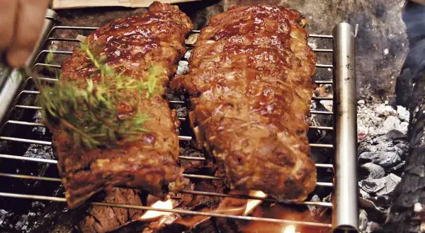 Två saftiga spareribs grillas över öppen eld. En hand lägger på en kvist rosmarin för extra smak.