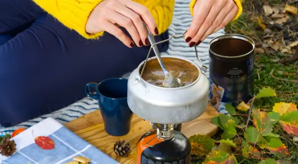 Preparando café en la naturaleza con recetas camping gas. Una tetera hierve sobre un hornillo, junto a una taza azul y galletas.